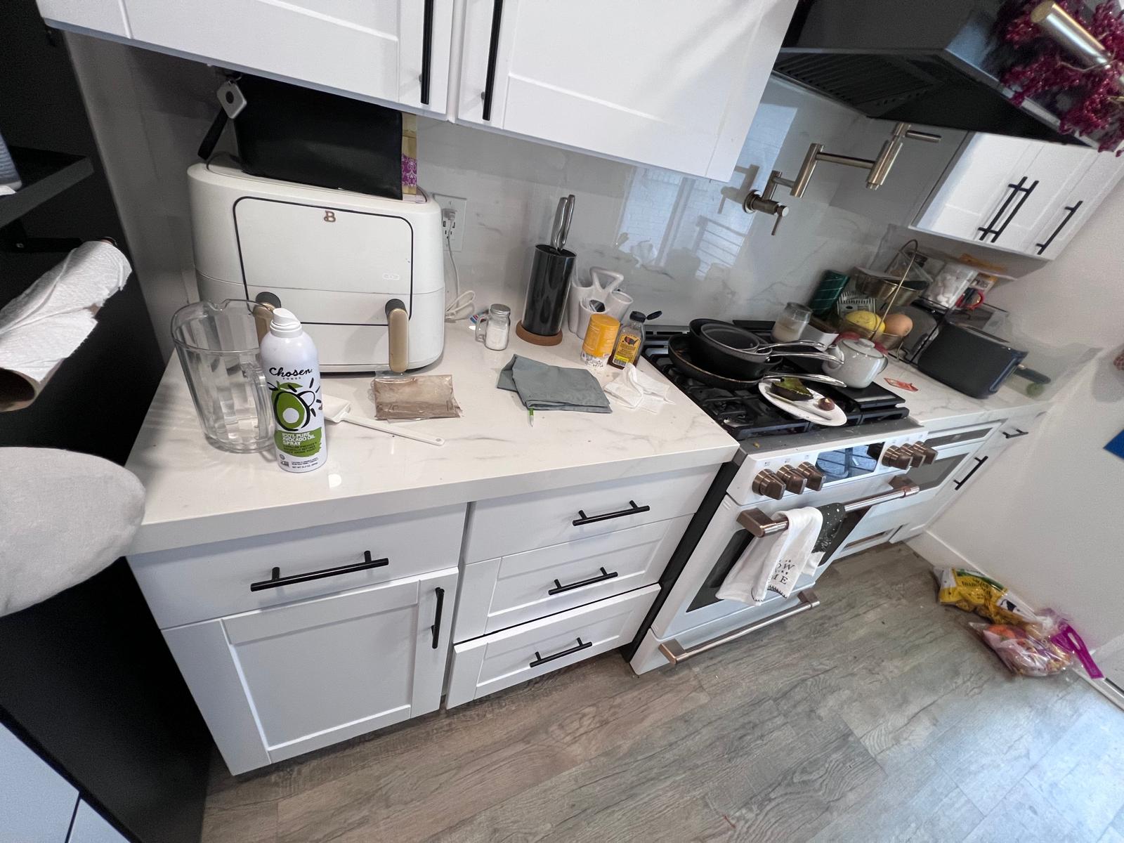Residential kitchen before cleaning with cluttered counters and floor items.